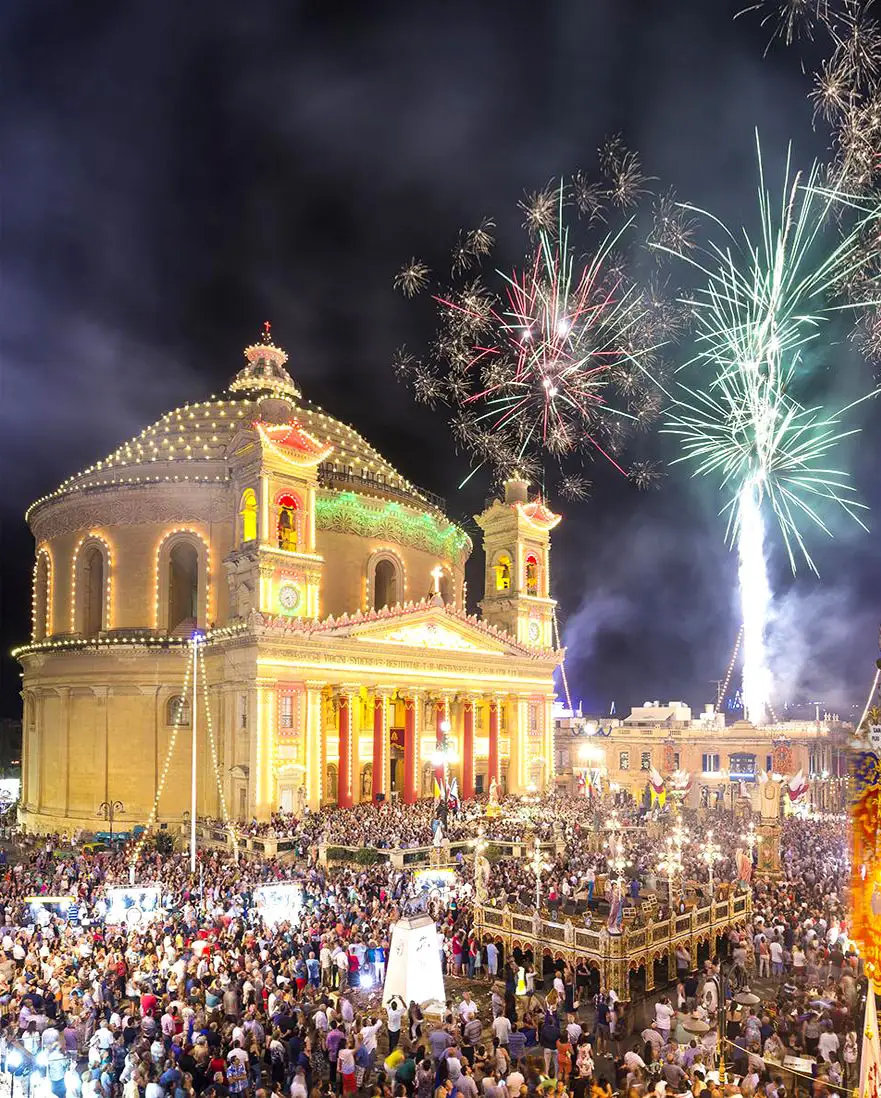 Malta village festa celebration with illuminated church, fireworks display, large crowd, and traditional summer festival atmosphere in Mediterranean Europe